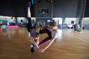 2 women hold hands 1 upside down as they navigate dancing dancing on harness in counterbalance.