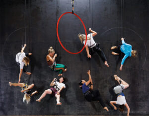 Against a black wall, 4 pairs of dancers chat whilst hanging in harness. A red hoop hangs in the foreground. 