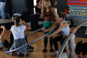 A female instructor shares a joke with 3 participants who stand, sit & kneel within a large metal (cyr) wheel. 