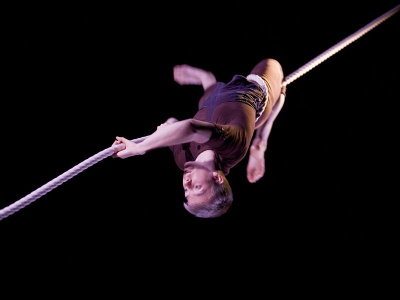 An aerialist is suspended upside down with a corde lisse entwined around their hips and knee, looking down the rope expectantly