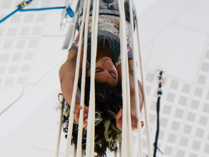 Aerialist Shereen Hussain hangs upside down surrounded by many cords of rope, arms hang relaxed overhead toward the floor, eyes closed