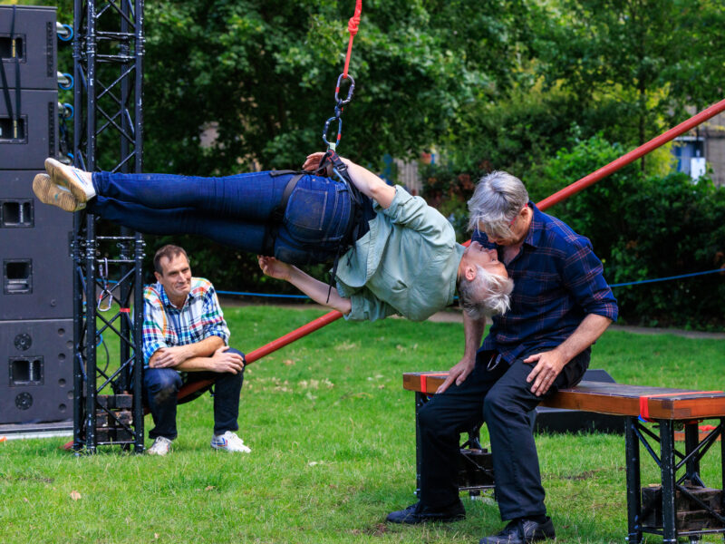 A man seated on a bench gazes down into his dance partners face as she flies, body held in a horizontal position supported by rope and harness. Behind them seated on a diagonal pole another male dancer laughs with them